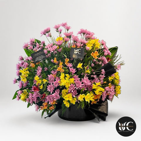 Large rounded floral arrangement in a black bowl with abundant greenery and pink, yellow, and orange flowers displayed on a counter in a flower shop.