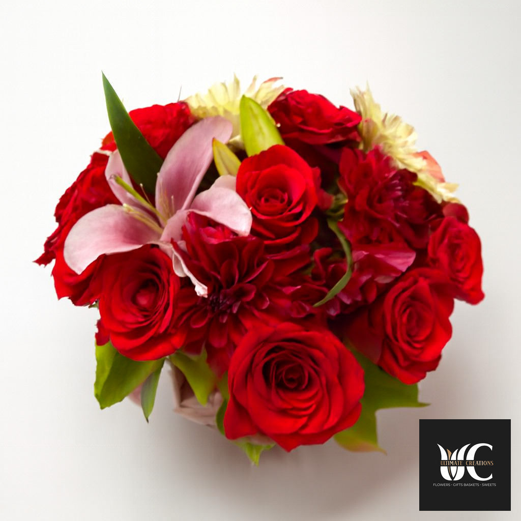 Round bouquet of red roses and mixed pink and cream flowers with green leaves on a white background.