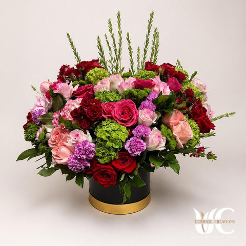 Full flower-box arrangement with red, pink, peach, lavender, and green flowers in a black container with a gold rim.