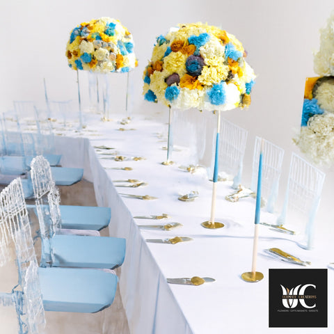 Long banquet table with tall dome floral centerpieces in blue, yellow, and cream, white linens, gold accents, and light blue chairs.