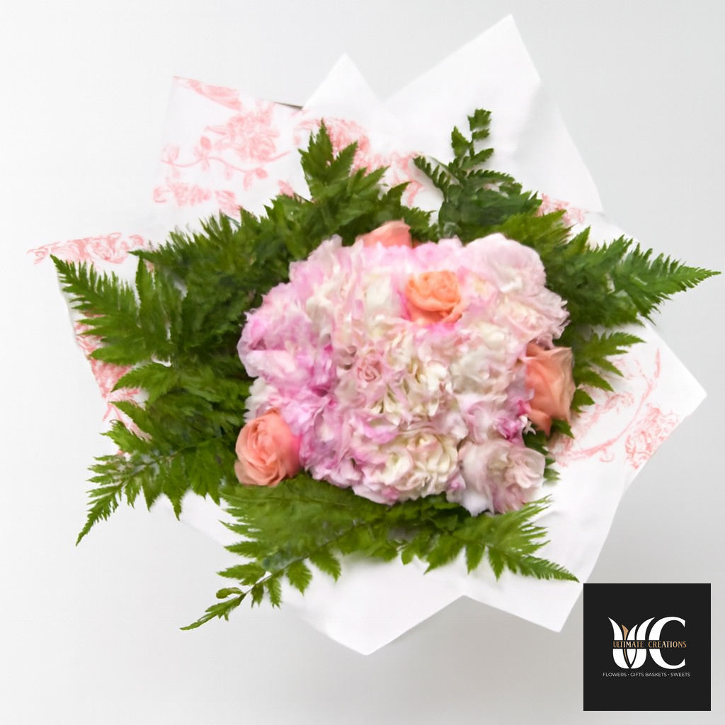 Bouquet of pink and white flowers with peach roses and green fern-like foliage, wrapped in white patterned paper.