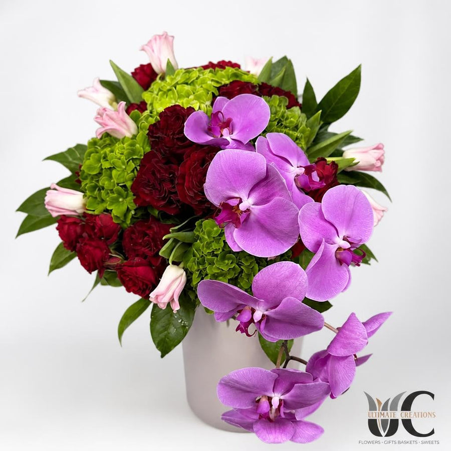Lush square vase arrangement with purple orchids, burgundy roses, green blooms, pink accents, and foliage