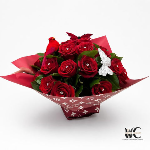 Bouquet of deep red roses with green leaves in red patterned wrapping, finished with a silver bow and a small red decorative accent, resting on a white table.