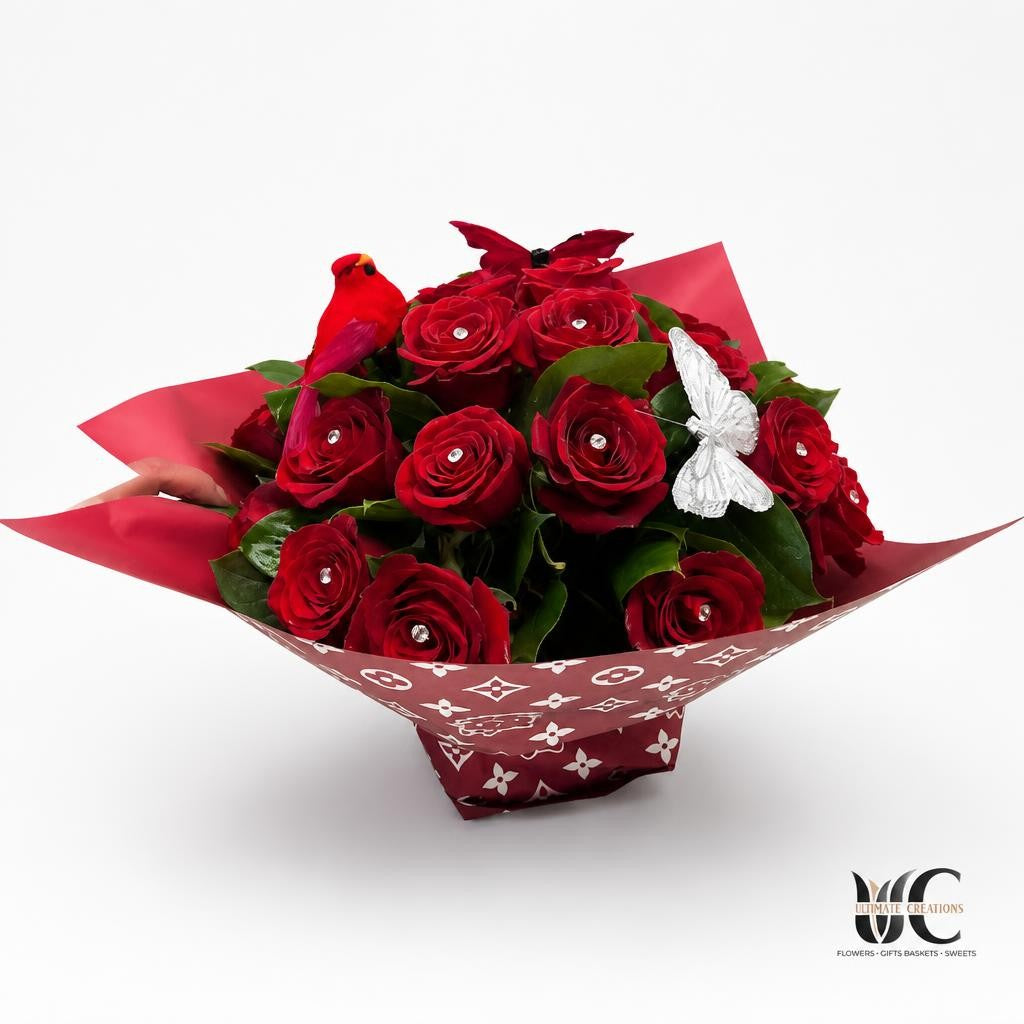 Bouquet of deep red roses with green leaves in red patterned wrapping, finished with a silver bow and a small red decorative accent, resting on a white table.